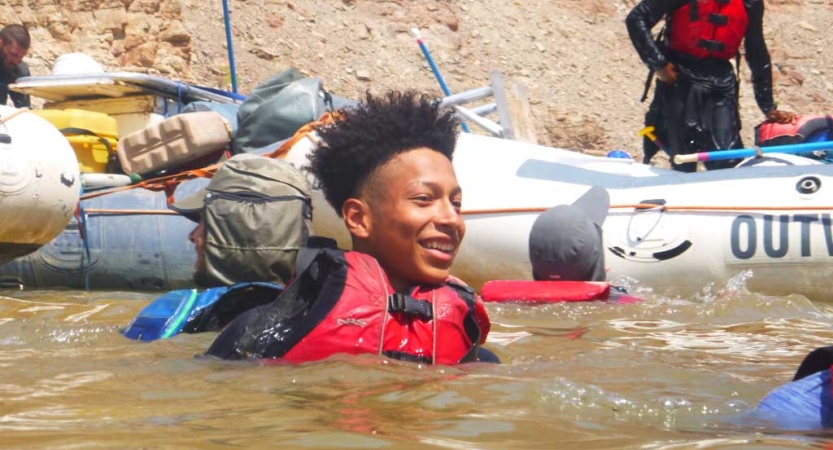 A person wearing a lifejackets smiles as they float in water. There is a raft behind them. 